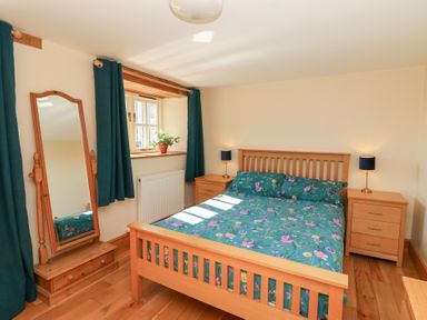 A bedroom with a wooden bed with floral bedding two bedside tables with lamps a standing mirror a window with curtains and a plant on the windowsill at The Stable in Aberaeron