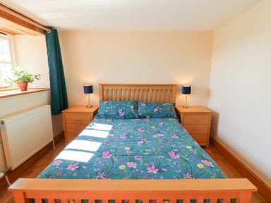 A bedroom with a wooden bed with floral bedding two bedside tables with lamps and a window with a plant at The Stable in Aberaeron