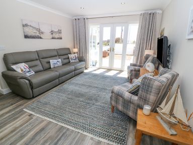 A living room with a leather sofa, two plaid armchairs, a wooden side table with a model sailboat and candles, a wall-mounted TV, and French doors with curtains at St Winifreds in Rhosneigr