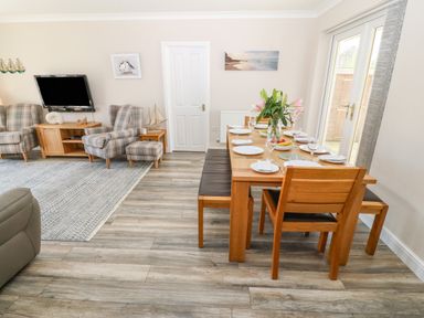 A dining room with a wooden table set with plates and glasses next to a living area with plaid armchairs and a wall mounted television at St Winifreds in Rhosneigr