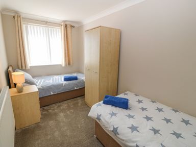A bedroom with two single beds with star patterned bedding a wardrobe a bedside table with a lamp and a window with curtains at St Winifreds in Rhosneigr