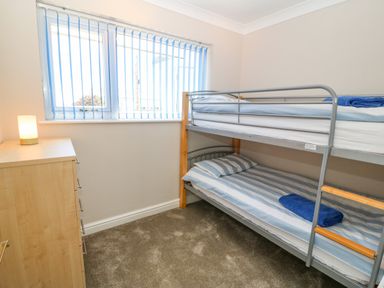 A small bedroom with a bunk bed a wooden dresser with a lamp and a window with vertical blinds at St Winifreds in Rhosneigr