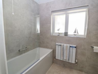 A bathroom with a bathtub and glass screen a window with decorative items a radiator with a towel and tiled walls at St Winifreds in Rhosneigr