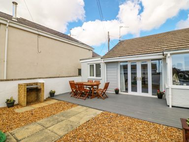 An outdoor patio area with a wooden dining table and chairs a small brick barbecue and gravel ground at St Winifreds Rhosneigr