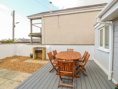 An outdoor patio area with a wooden dining table and six chairs a built-in brick barbecue and a pebble and stone ground surface at St Winifreds in Rhosneigr