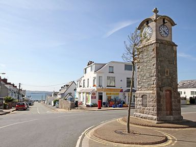 A stone clock tower and a small tree on a street corner with houses and shops in the background at St Winifreds in Rhosneigr