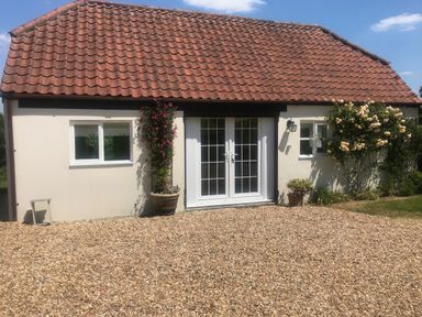 A single story building with a tiled roof two windows and a double glass door with plants and flowers outside at Oke Apple Cottage in Okeford Fitzpaine