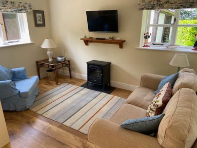 A living room with a beige sofa a blue armchair a striped rug a wall-mounted TV and a small stove at Oke Apple Cottage in Okeford Fitzpaine