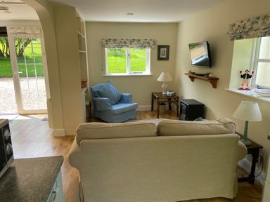 A living room with armchair sofa lamps television and windows showing grass at Oke Apple Cottage in Okeford Fitzpaine