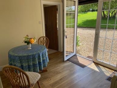 A dining area with a round table covered with a blue tablecloth and wooden chairs near glass doors opening to a garden at Oke Apple Cottage in Okeford Fitzpaine