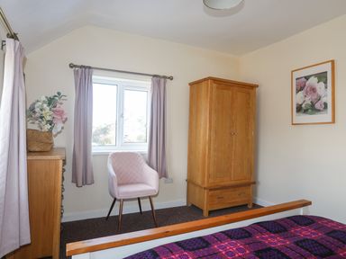 A bedroom with a wooden wardrobe a pink chair a wooden dresser with flowers on top and a bed with a purple checkered blanket at Glan Y Don Bach in Abersoch