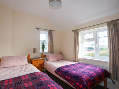 A bedroom with two single beds covered with pink sheets and purple blankets a wooden nightstand with a lamp between the beds and beige curtains on windows at Glan Y Don Bach in Abersoch