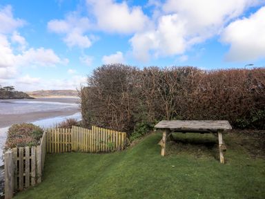 A garden area with a wooden picnic table and wooden fence overlooking a beach and water at Glan Y Don Bach in Abersoch
