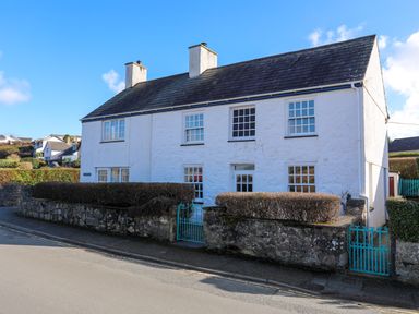 A white stone house with multiple windows and two chimneys behind a low stone wall with blue gates at Glan Y Don Bach in Abersoch