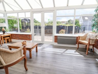 A sunroom with large windows two wicker chairs a wooden coffee table and a small table with a lamp at Beach Cottage in Conwy