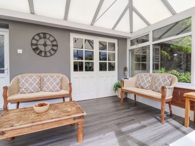 A conservatory with two wicker benches, a wooden coffee table, a wall clock, and large windows at Beach Cottage in Conwy