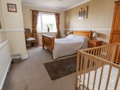 A bedroom with a wooden bed a crib a wooden wardrobe a nightstand with a lamp a chair with towels and a window with curtains at Beach Cottage in Conwy