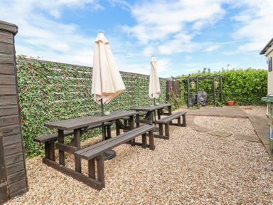 An outdoor patio area with two picnic tables and umbrellas on a gravel surface with a barbecue grill and hedges in the background at Beach Cottage in Conwy