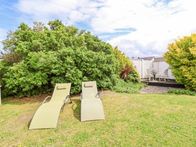 An outdoor garden with two lounge chairs on the grass and a small patio table with chairs behind bushes at Beach Cottage in Conwy