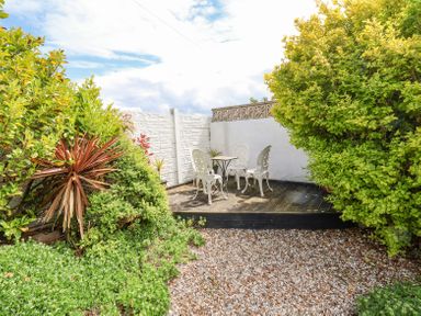 A small outdoor patio area with three white metal chairs and a table surrounded by bushes and a gravel ground at Beach Cottage in Conwy