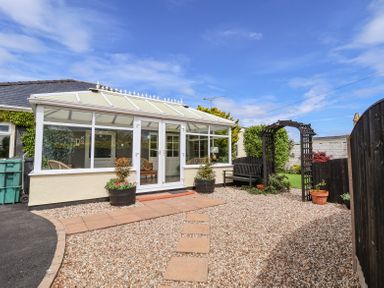 A conservatory with glass windows and doors leading to a gravel garden with stepping stones and a wooden bench under an arch at Beach Cottage in Conwy