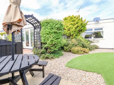 A garden area with a wooden picnic table and umbrella near a pathway bordered by gravel and a lawn at Beach Cottage in Conwy