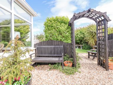 A garden area with a wooden bench next to a trellis archway and gravel path outside a conservatory at Beach Cottage in Conwy