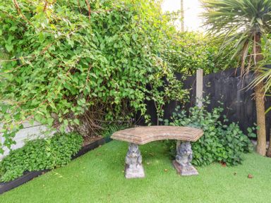 A garden with a curved stone bench supported by two animal-shaped legs on artificial grass surrounded by plants and a black fence