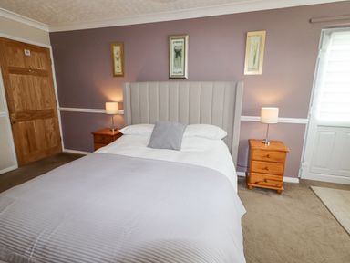 A bedroom with a double bed a gray headboard two wooden nightstands with lamps and floral artwork on wall at Beach Cottage in Conwy
