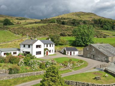A farmhouse and outbuildings with garden and hills in the background at Ghyll Bank House in Staveley near Windermere