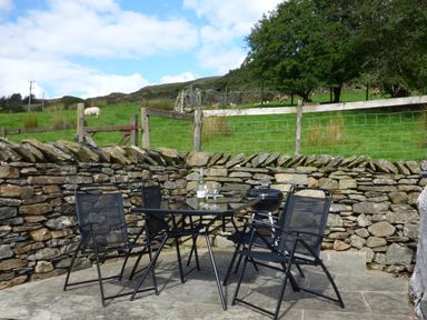 An outdoor patio with a glass table and four black mesh chairs next to a stone wall and green fields at Ghyll Bank Bungalow in Staveley near Windermere