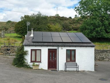 A small white house with solar panels on the roof a red door and two windows with vertical blinds at Ghyll Bank Bungalow in Staveley near Windermere
