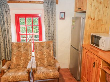Two patterned armchairs in front of a red window with curtains beside a refrigerator and wooden kitchen cabinets at Willowbrook Cottage in Askill County Leitrim