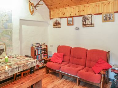 A room with a red three-seater sofa and pillows a small bookshelf filled with books a table with a tablecloth and placemats wooden ceiling and framed pictures on the wall at Willowbrook Cottage in Askill County Leitrim
