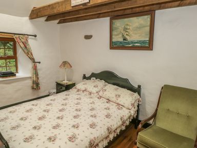 A bedroom with a floral bedspread on a double bed a green upholstered chair a small bedside table with a lamp curtains on a window and a painting of a sailing ship on the wall at Willowbrook Cottage in Askill County Leitrim