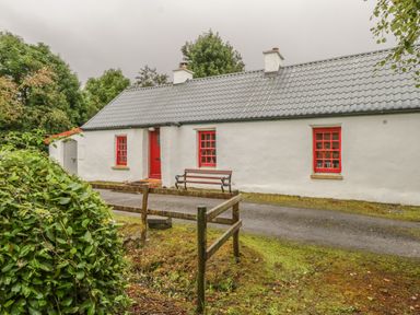 A white cottage with a gray roof red door and red window frames a bench outside the house on a gravel driveway with greenery around at Willowbrook Cottage in Askill County Leitrim