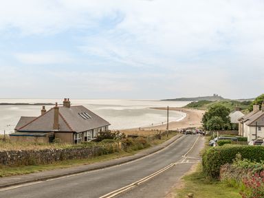 A coastal road leading to a beach with houses on both sides and a castle in the distance at Coastguard Cottage in Low Newton-by-the-Sea near Beadnell