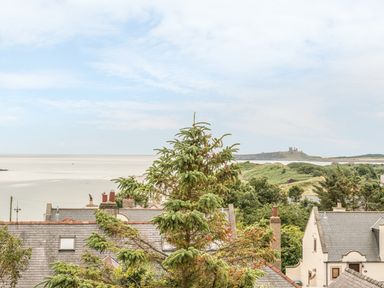 A coastal view with rooftops and trees overlooking a beach and distant castle at Coastguard Cottage in Low Newton-by-the-Sea near Beadnell