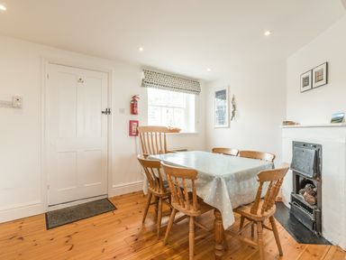 A dining area with a wooden table covered by a polka dot tablecloth and six wooden chairs next to a window and a fireplace at Coastguard Cottage in Low Newton-by-the-Sea near Beadnell
