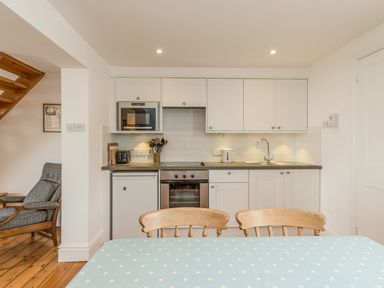 A kitchen with white cabinets, an oven, microwave, toaster, kettle, and sink with wooden chairs and a table with a blue polka dot tablecloth at Coastguard Cottage in Low Newton-by-the-Sea near Beadnell