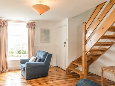 A room with a blue armchair near a window with striped curtains and wooden stairs at Coastguard Cottage in Low Newton-by-the-Sea near Beadnell