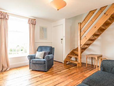 A living room with a blue armchair a wooden staircase and small tables at Coastguard Cottage Low Newton-by-the-Sea near Beadnell