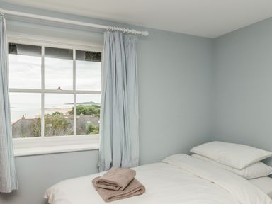 A bedroom with a single bed with white bedding and folded towels near a window with curtains overlooking rooftops and a distant coast at Coastguard Cottage in Low Newton-by-the-Sea near Beadnell