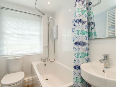 A bathroom with a bathtub shower combo a fish patterned shower curtain a toilet a window with blinds and a sink at Coastguard Cottage in Low Newton-by-the-Sea near Beadnell