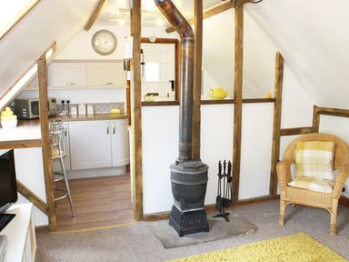 A room with a wood stove a wicker chair and a kitchen area at The Hayloft in Necton near Swaffham