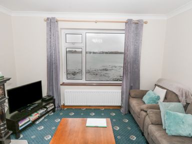 A living room with a sofa a wooden coffee table a television on a stand and a large window overlooking water at Quayside House in Roundstone County Galway