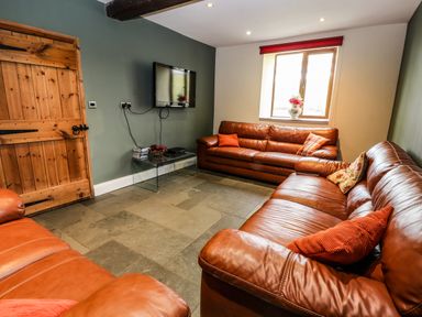 A sitting room with two brown leather sofas a wooden door a wall-mounted TV and a window with a vase of flowers at Maestyddyn Farm House in Clawddnewydd near Ruthin
