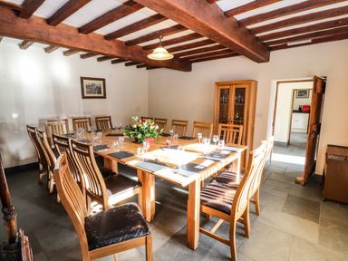 A dining room with a large wooden table set with glasses and placemats surrounded by wooden chairs with a flower arrangement on the table at Maestyddyn Farm House in Clawddnewydd near Ruthin