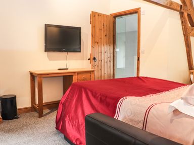 A bedroom with a red bedspread wooden desk wall-mounted tv and open wooden door at Maestyddyn Farm House Clawddnewydd near Ruthin