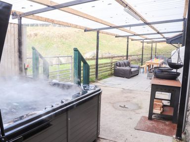 An outdoor covered patio with a steaming hot tub a leather sofa a wooden table with colorful cups and a wood stove at Maestyddyn Farm House Clawddnewydd near Ruthin